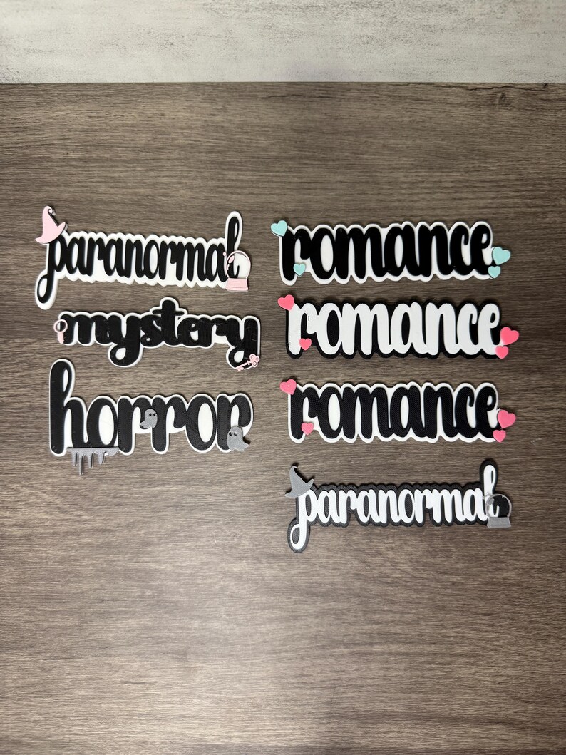 Puede incluir: Palabras troqueladas en blanco y negro, incluyendo "paranormal", "romance", "misterio" y "horror". Algunas palabras tienen elementos decorativos como corazones, fantasmas y sombreros de bruja. Los art&iacute;culos est&aacute;n dispuestos sobre una superficie de madera.