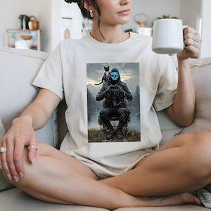 May include: Cream-colored t-shirt featuring a graphic of a figure with a blue skull face, seated with a black cat on their shoulder. The figure is making a heart shape with their hands. The background shows a forest and lake.