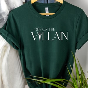 May include: Dark green t-shirt with rolled-up sleeves. The shirt features the text "Dibs on the Villain" in white, with a sword graphic. The shirt is hanging on a wooden hanger.