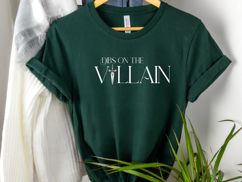 May include: Dark green t-shirt with rolled-up sleeves. The shirt features the text "Dibs on the Villain" in white, with a sword graphic. The shirt is hanging on a wooden hanger.
