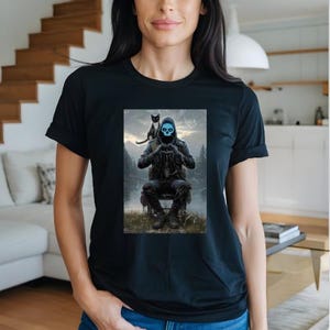 May include: Black t-shirt featuring a graphic of a hooded figure with a blue skull face, seated with a black cat on their shoulder. The artwork depicts a forest scene with a lake and mountains in the background. The t-shirt is made of a soft material.