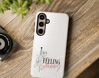 Peut inclure: Coque de téléphone blanche avec un contour noir d'un poignard et de fleurs. Le texte "Feeling Stabby" est écrit en rouge.