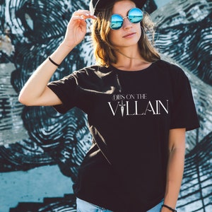 May include: Black t-shirt with the text "Dibs on the Villain" in white. The shirt is a crew neck style and is being worn by a person. The background is a graffiti wall with black and blue paint.