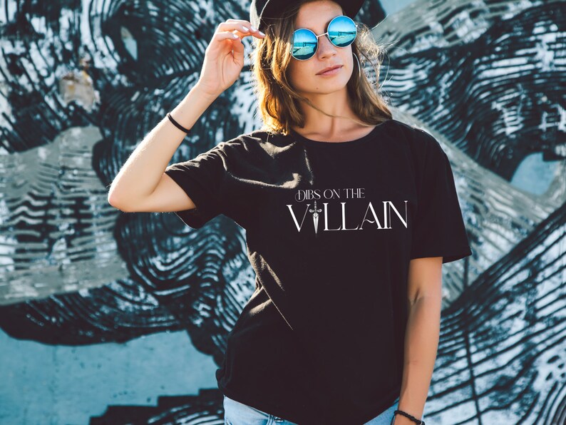 May include: Black t-shirt with the text "Dibs on the Villain" in white. The shirt is a crew neck style and is being worn by a person. The background is a graffiti wall with black and blue paint.