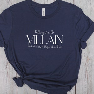 K&ouml;nnte beinhalten: Marineblaues T-Shirt mit dem wei&szlig;en Text "Falling for the VILLAIN One Page at a Time". Das T-Shirt hat gerollte &Auml;rmel und einen geknoteten Saum. Das Design beinhaltet eine kleine Buchillustration. Das T-Shirt ist aus weichem Material.