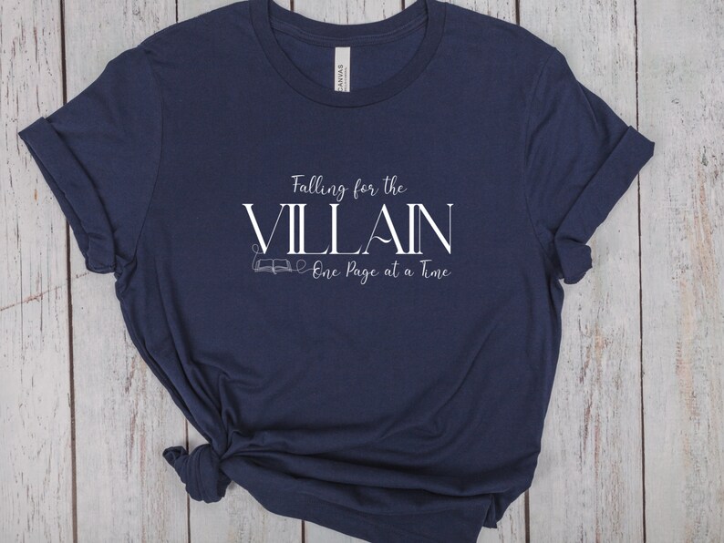 K&ouml;nnte beinhalten: Marineblaues T-Shirt mit dem wei&szlig;en Text "Falling for the VILLAIN One Page at a Time". Das T-Shirt hat gerollte &Auml;rmel und einen geknoteten Saum. Das Design beinhaltet eine kleine Buchillustration. Das T-Shirt ist aus weichem Material.