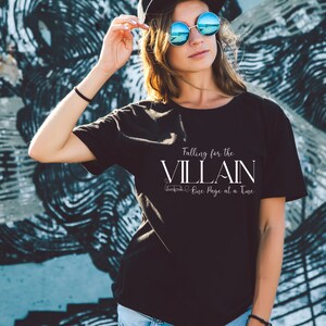 K&ouml;nnte beinhalten: Schwarzes T-Shirt mit dem wei&szlig;en Text "Falling for the VILLAIN One Page at a Time". Das T-Shirt hat einen Rundhalsausschnitt und kurze &Auml;rmel. Der Hintergrund zeigt eine blau-graue Graffiti-Wand.