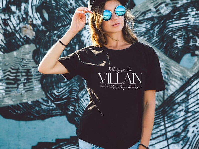 K&ouml;nnte beinhalten: Schwarzes T-Shirt mit dem wei&szlig;en Text "Falling for the VILLAIN One Page at a Time". Das T-Shirt hat einen Rundhalsausschnitt und kurze &Auml;rmel. Der Hintergrund zeigt eine blau-graue Graffiti-Wand.
