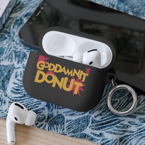 May include: A black silicone protective case for wireless earbuds featuring the yellow text "GODDAMNIT DONUT" with a small red crown icon above the text and a red starburst graphic behind the word "DONUT". Includes a silver metal keyring attachment.
