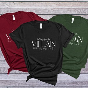 K&ouml;nnte beinhalten: Drei T-Shirts in Burgunderrot, Schwarz und Tannengr&uuml;n. Jedes Shirt tr&auml;gt den wei&szlig;en Text "Falling for the Villain One Page at a Time" mit einer Buchillustration. Die Shirts sind unten geknotet.
