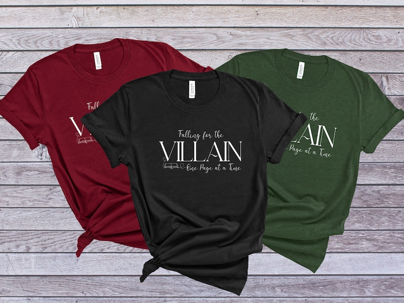 K&ouml;nnte beinhalten: Drei T-Shirts in Burgunderrot, Schwarz und Tannengr&uuml;n. Jedes Shirt tr&auml;gt den wei&szlig;en Text "Falling for the Villain One Page at a Time" mit einer Buchillustration. Die Shirts sind unten geknotet.