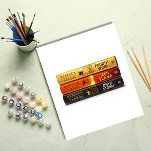 May include: A paint-by-numbers kit featuring a printed illustration of three stacked book spines by Rebecca Yarros titled Fourth Wing, Iron Flame, and Onyx Storm. The set includes a cup of paintbrushes, three additional brushes, and small numbered paint pots.