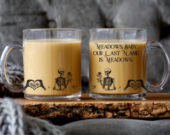Può includere: Due tazze in vetro trasparente riempite con una bevanda marrone chiaro. Ogni tazza presenta una grafica nera di uno scheletro che tiene una rosa e mani che formano un cuore. La tazza a destra riporta il testo: MEADOWS, BABY. OUR LAST NAME IS MEADOWS.