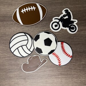 Puede incluir: Colecci&oacute;n de elementos decorativos con tem&aacute;tica deportiva. Incluye un bal&oacute;n de f&uacute;tbol americano marr&oacute;n con rayas blancas, una moto negra con un piloto, un voleibol blanco y negro, un bal&oacute;n de f&uacute;tbol negro y blanco, una pelota de b&eacute;isbol con costuras rojas y un sombrero de vaquero marr&oacute;n.
