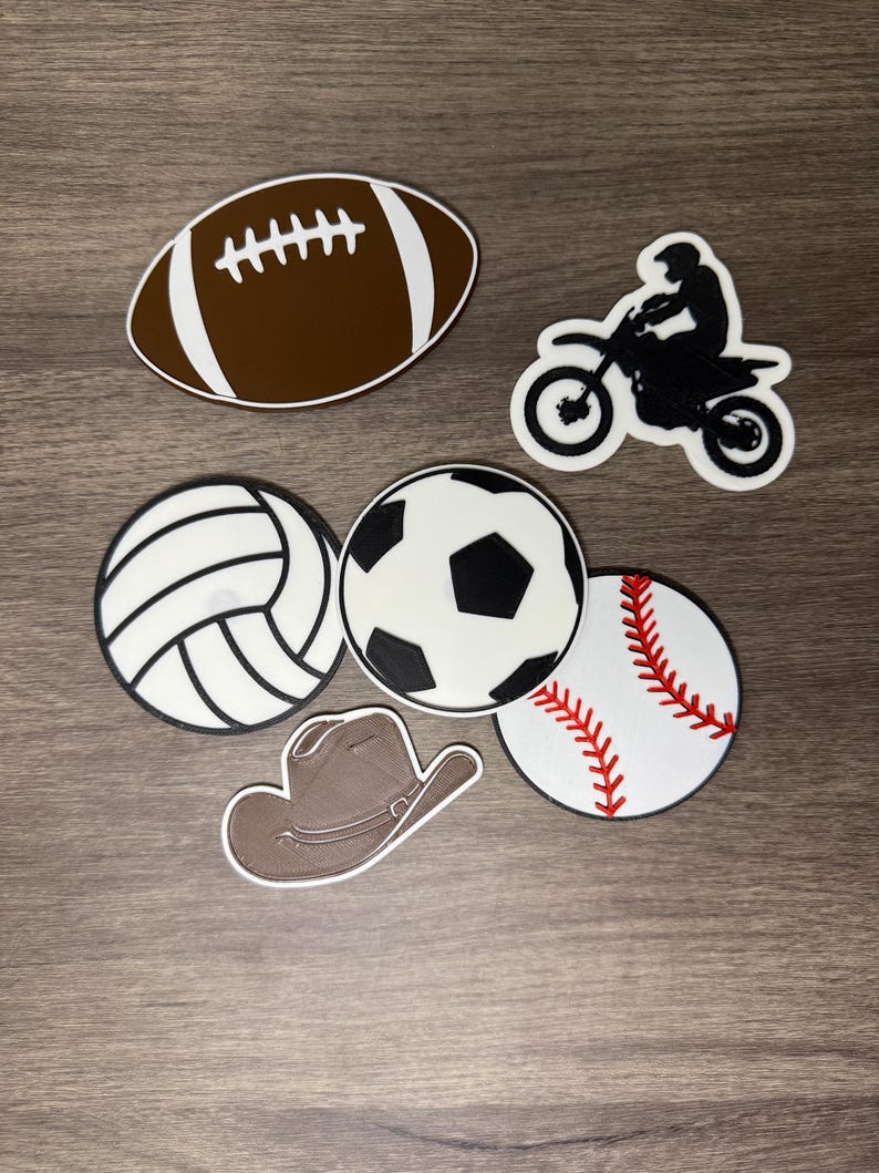 Puede incluir: Colecci&oacute;n de elementos decorativos con tem&aacute;tica deportiva. Incluye un bal&oacute;n de f&uacute;tbol americano marr&oacute;n con rayas blancas, una moto negra con un piloto, un voleibol blanco y negro, un bal&oacute;n de f&uacute;tbol negro y blanco, una pelota de b&eacute;isbol con costuras rojas y un sombrero de vaquero marr&oacute;n.
