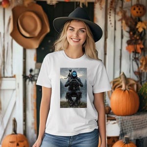 May include: White t-shirt featuring a graphic of a person with a blue skull mask and a black cat on their shoulder. The graphic is set against a misty, outdoor backdrop. The woman is wearing a black hat.
