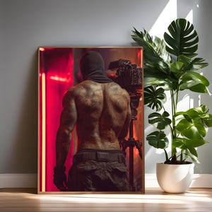 Op de afbeelding: Ingelijste kunstafdruk met een gespierde figuur met tatoeages, gekleed in een zwarte bivakmuts en een camera op een statief vasthoudend. De achtergrond heeft een rood en zwart kleurenschema. Een grote potplant staat naast de ingelijste kunst.