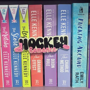 May include: A shelf of books with colorful spines, including titles like "The Mistake," "The Score," and "The Legacy." A decorative sign that says "Hockey Romance" is in front of the books. The books are by Elle Kennedy and Emily Rath.