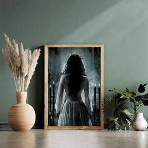 May include: A framed print of a woman in a white dress standing in a dark, shadowy room. The woman's back is to the viewer, and she has long, dark hair. There are candles on either side of her.