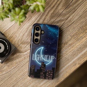 Puede incluir: Una funda de teléfono negra con un diseño de cielo nocturno estrellado azul y blanco. La funda presenta una luna creciente de neón con el texto "LIGHT IT UP" en su interior. La funda también presenta una silueta de un horizonte de ciudad.