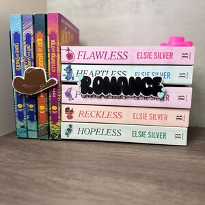 Puede incluir: Una pila de novelas rom&aacute;nticas de Elsie Silver con t&iacute;tulos como "Flawless" y "Heartless". Se ve un marcap&aacute;ginas con forma de sombrero de vaquero marr&oacute;n y un tope de libros con forma de sombrero de vaquero rosa. La palabra "Romance" est&aacute; escrita en letras negras.