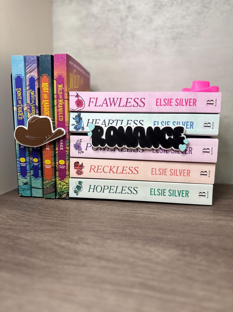 Puede incluir: Una pila de novelas rom&aacute;nticas de Elsie Silver con t&iacute;tulos como "Flawless" y "Heartless". Se ve un marcap&aacute;ginas con forma de sombrero de vaquero marr&oacute;n y un tope de libros con forma de sombrero de vaquero rosa. La palabra "Romance" est&aacute; escrita en letras negras.