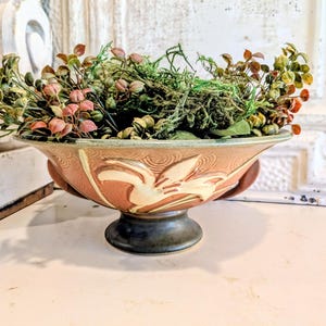 Può includere: Una ciotola in ceramica su piedistallo con esterno color terracotta, motivi floreali in rilievo color crema e una base marrone scuro. La ciotola è riempita con una varietà di fogliame artificiale verde e marrone rossastro.