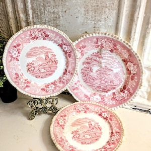 May include: Three ceramic plates featuring a pink and white transferware pattern. The design includes detailed pastoral landscapes with buildings and grazing animals in the center, surrounded by a border of floral motifs and scalloped edges.