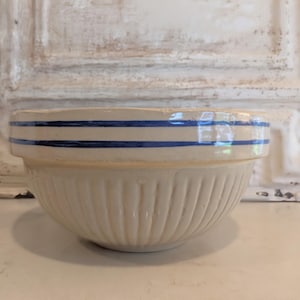 May include: A cream-colored ceramic mixing bowl featuring a smooth upper rim decorated with two horizontal blue stripes. The lower portion of the bowl has a vertical ribbed texture, providing a distinct contrast to the plain, smooth upper section.