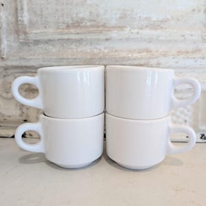 May include: Four small, white ceramic coffee cups are stacked in two pairs. Each cup has a rounded handle and a simple, minimalist design. The cups are set against a neutral, textured background.