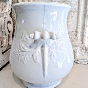 May include: A white ceramic vessel featuring a wide, flared rim and a rounded body. The exterior is decorated with embossed leaf patterns and a central handle shaped like a stylized scroll. The surface has a glossy finish with visible glaze.