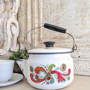 Puede incluir: Tetera de esmalte blanco con asa y tapa negras. La tetera está decorada con un colorido diseño floral en rojo, naranja, verde y azul. El asa es negra y el pico está en el lado izquierdo. La tetera está sobre una superficie blanca.