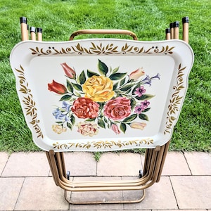 May include: A set of four folding snack trays with a cream-colored surface featuring a central floral bouquet of yellow, red, and pink roses with green leaves. The trays have gold-toned metal frames and decorative gold leaf-patterned borders.