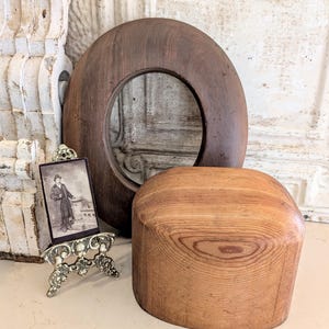 May include: A wooden hat form and a circular wooden object, possibly a hat stretcher, sit on a light-colored surface. A small framed photograph in a decorative stand is also present. The wood tones range from light to dark brown.