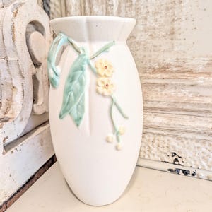 May include: A white ceramic vase with a subtle crackle glaze finish, featuring a raised relief design of pale green leaves and small yellow flowers on a vine. The vase has a tapered oval shape with a flared rim and a single handle on the side.