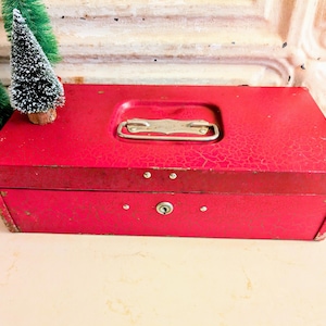 May include: A rectangular red metal lockbox featuring a weathered, crackled paint finish, a central silver-toned metal handle, and a keyhole on the front panel. Two small, green, snow-dusted artificial pine trees are positioned to the left side.