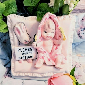 Könnte beinhalten: Rosa Keramik-Babyfigur, eingewickelt in ein gepunktetes Handtuch, hält einen Spiegel und einen Kamm. Ein Hasenkopf und ein Schild mit der Aufschrift "PLEASE DON'T DISTURB" sind ebenfalls abgebildet. Die Figur steht auf einem kissenförmigen Sockel.