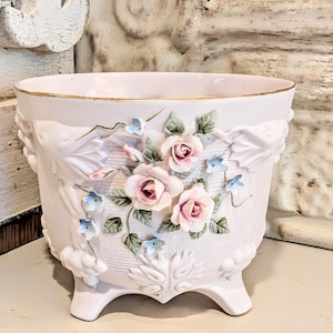 May include: A pale pink ceramic planter featuring a gold-trimmed rim and three feet. The exterior is decorated with raised, three-dimensional light pink roses, green leaves, and small blue flowers against a textured, basket-weave patterned surface.