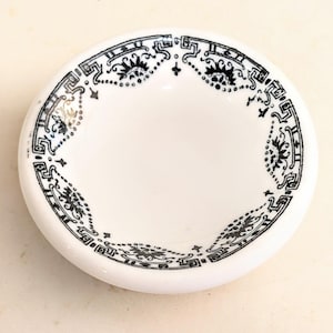 Puede incluir: Un pequeño plato redondo de cerámica blanca con un patrón decorativo negro en el borde interior. El diseño consiste en formas geométricas repetitivas, motivos florales y líneas punteadas que crean un marco continuo alrededor.