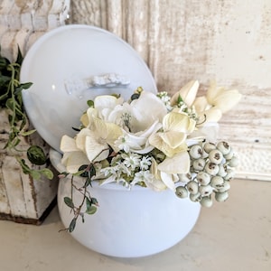 May include: A light blue, round ceramic container with a lid, filled with white and cream-colored flowers and berries. The container has a handle on the lid and is sitting on a white surface. The flowers are arranged to spill over the edge of the container.