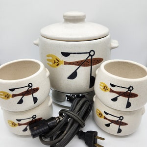May include: A vintage cream-colored fondue set with a matching lid, pot, and four bowls. Each piece features a whimsical design of a yellow fork and black spoon. The set includes a heating element and a power cord.
