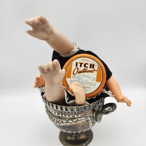 May include: A vintage silver cup with a doll inside, its arms and legs protruding. The doll is wearing a black dress with lace trim. A round orange and brown tin of "ITCH Ointment" is visible.