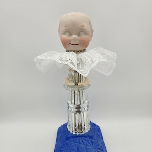 Könnte beinhalten: Eine dekorative Figur mit einem blassen, lächelnden Puppenkopf mit rosigen Wangen und geschlossenen Augen, der einen weißen Spitzenkragen trägt. Der Puppenkopf sitzt auf einer klaren, vertikal geriffelten Glassäule, die auf einem quadratischen kobaltblauen Glassockel montiert ist.