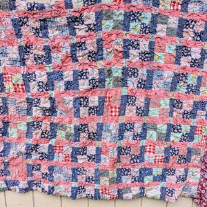 May include: A textured patchwork quilt featuring a grid of small fabric squares in various patterns, including floral, gingham, and solid prints. The design is dominated by alternating rows of dark blue and pink fabric strips, finished with a dark floral border.