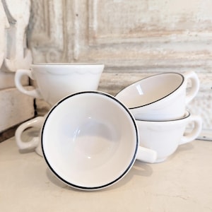 May include: A set of four white ceramic cups with a smooth finish and a thin black rim around the top edge. Each cup features a small handle on the side and a scalloped design detail along the upper exterior wall, arranged in a stacked cluster.