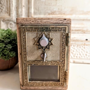 Puede incluir: Un buzón de madera de estilo vintage con una parte frontal de metal color latón. La parte frontal de metal presenta un diseño de estrella alrededor de un dial con letras y una pequeña ventana. El buzón tiene un ojo de cerradura y una puerta con bisagras.