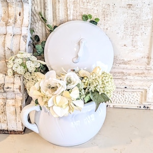 May include: A white ceramic container with a lid, filled with white and cream-colored flowers and greenery. The container has a handle and is set against a distressed white background, creating a rustic, farmhouse aesthetic.