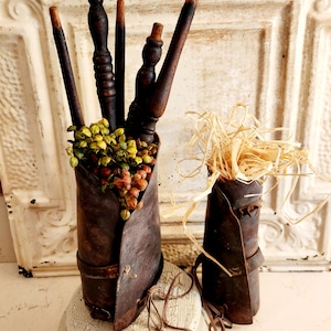 May include: Two rustic, dark brown metal containers with decorative wooden spindles and dried floral accents. One container holds several spindles and green and orange berries, while the other has straw. The containers sit on a distressed white pedestal.