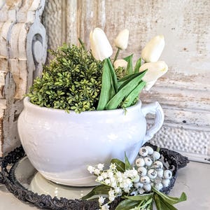 May include: A white ceramic teacup-shaped planter filled with green foliage and white tulips. The planter sits on a decorative black tray, with sprigs of white berries and lily of the valley. The background features distressed white wood.