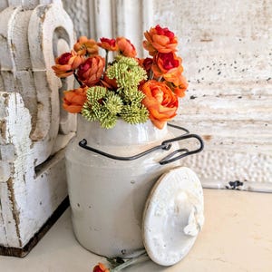 May include: A cream-colored ceramic milk jug with a black wire handle and a matching lid. The jug is filled with orange and red artificial flowers and green accents. The lid is resting beside the jug. The background is a distressed white surface.